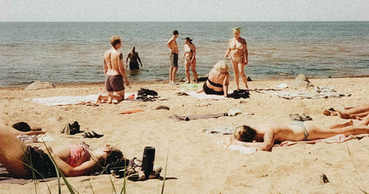 Sunbathers on a sandy beach on a bright summer day, some lying on towels and others standing at the water's edge