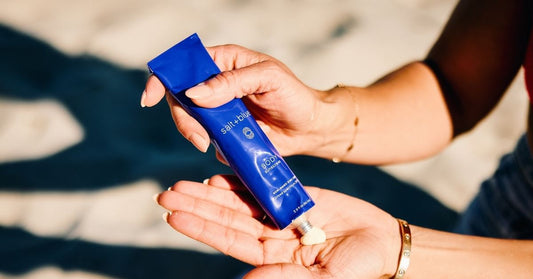 Hand squeezing salt+blue non-nano mineral sunscreen from a blue tube into a palm on the beach