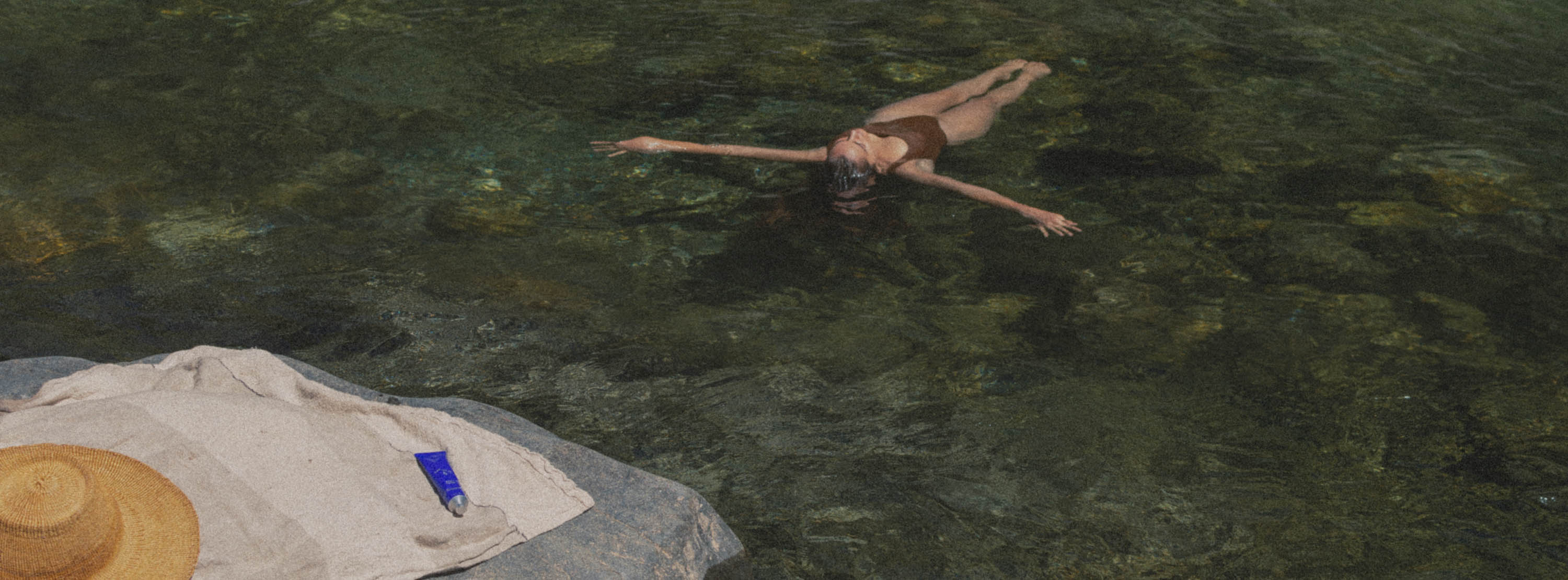 A woman swims in clear greenish water next to a rock with a white towel and a straw hat in the foreground.