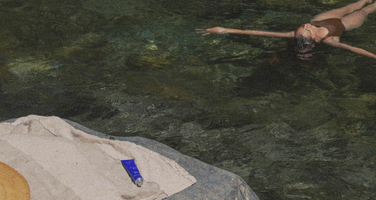 A woman swims in clear greenish water next to a stone or rock with a towel and a salt+blue BODY sunscreen.