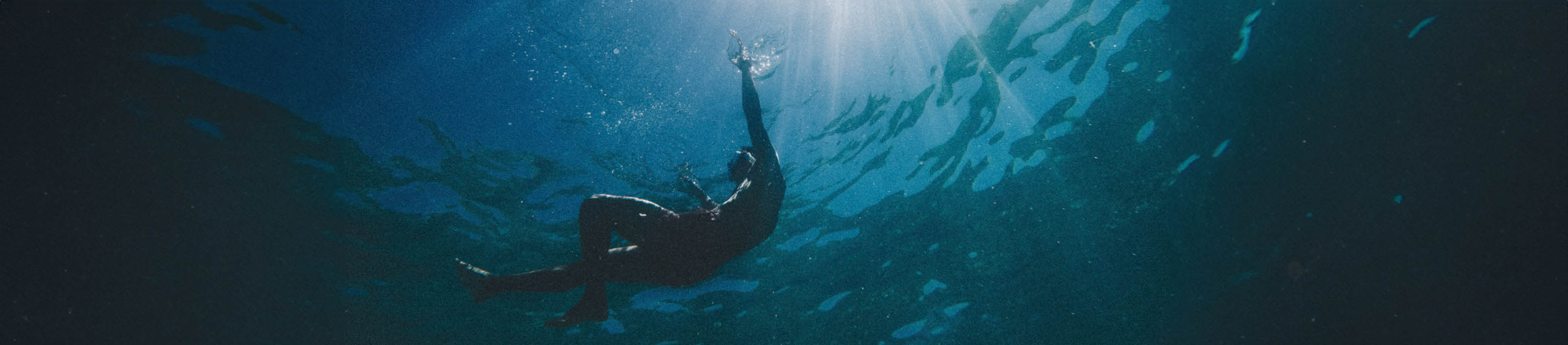 An underwater photograph of a diver swimming upwards toward bright sunbeams filtering through the water surface.