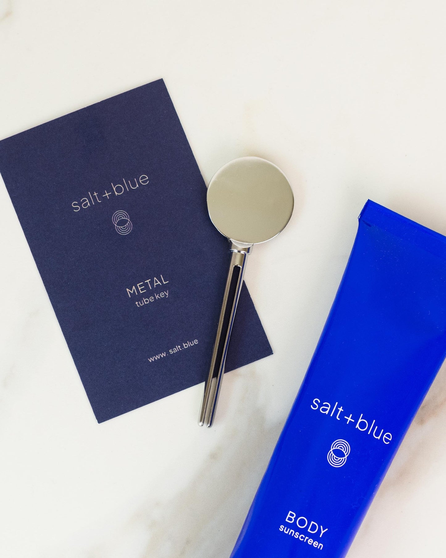 A flat-lay product shot of the salt+blue METAL Tub Key alongside its navy blue packaging card and bright blue body sunscreen tube, arranged on a white marble surface.
