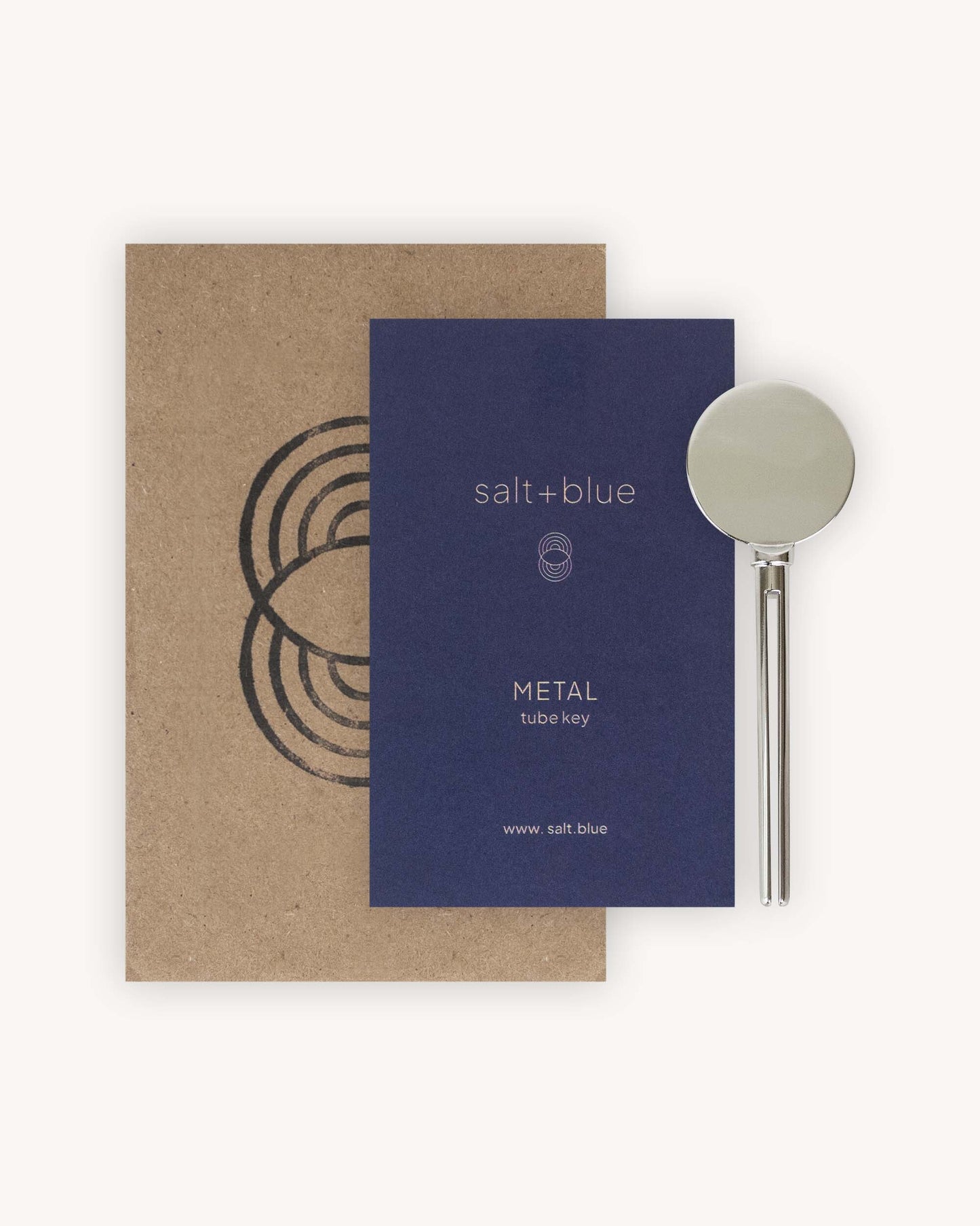 A salt+blue METAL tube key shown with its brown kraft paper envelope and a dark blue card insert on a light beige background.