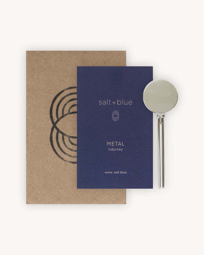 A salt+blue METAL tube key shown with its brown kraft paper envelope and a dark blue card insert on a light beige background.