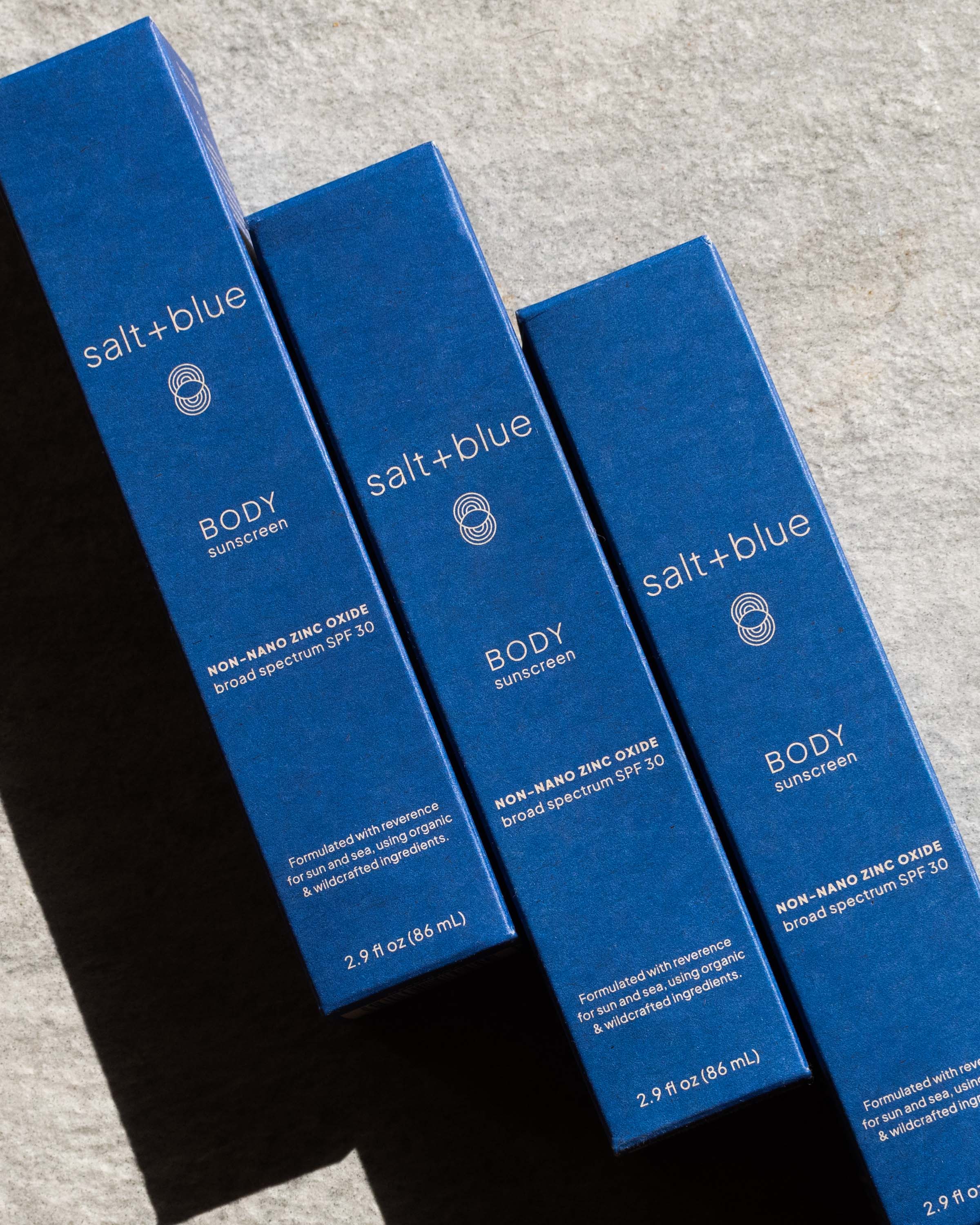 Three blue boxes of salt+blue body sunscreen on a gray surface