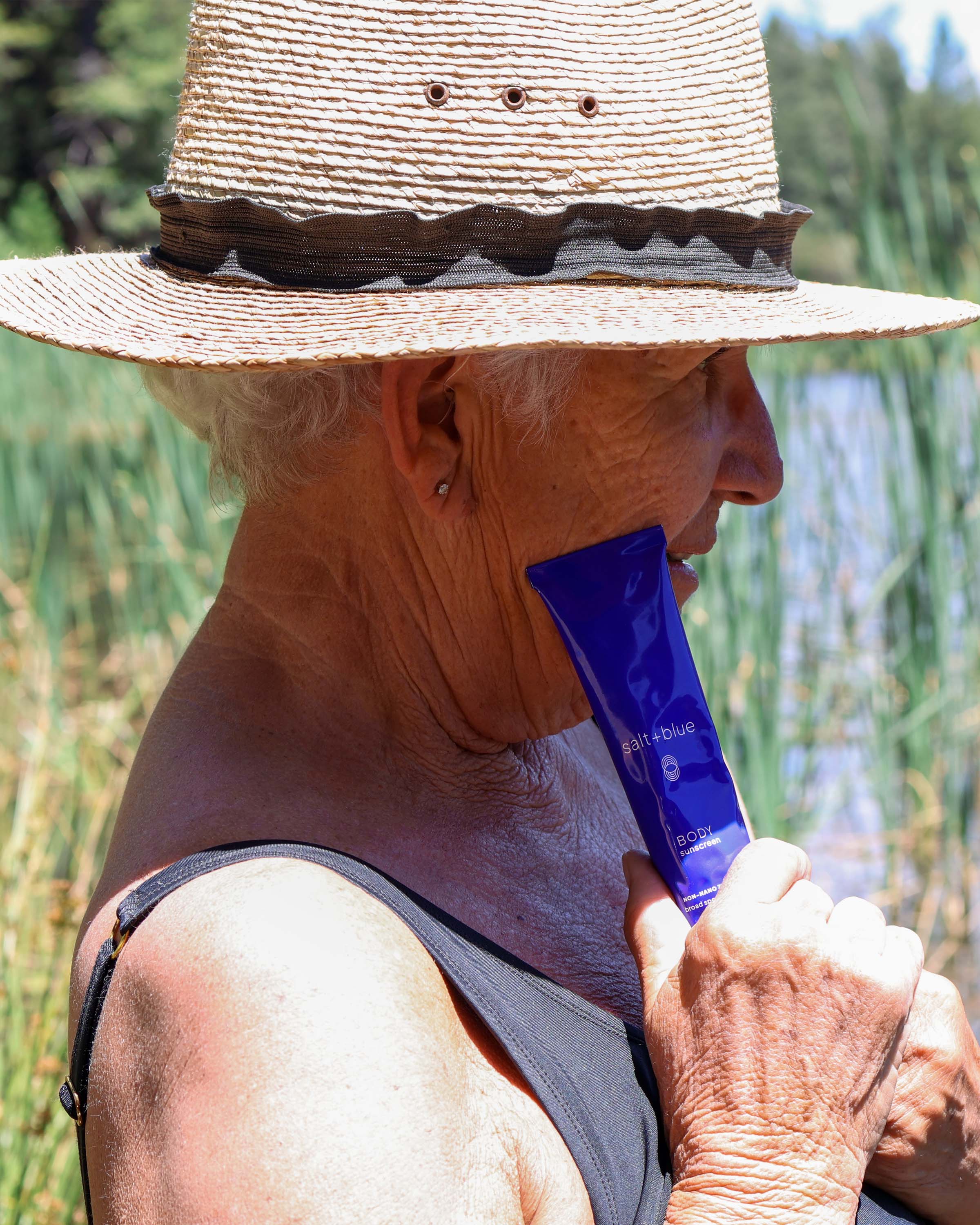 A woman in a straw sun hat and black tank top holds a blue salt+blue tube of BODY sunscreen near her face outdoors by a lake.