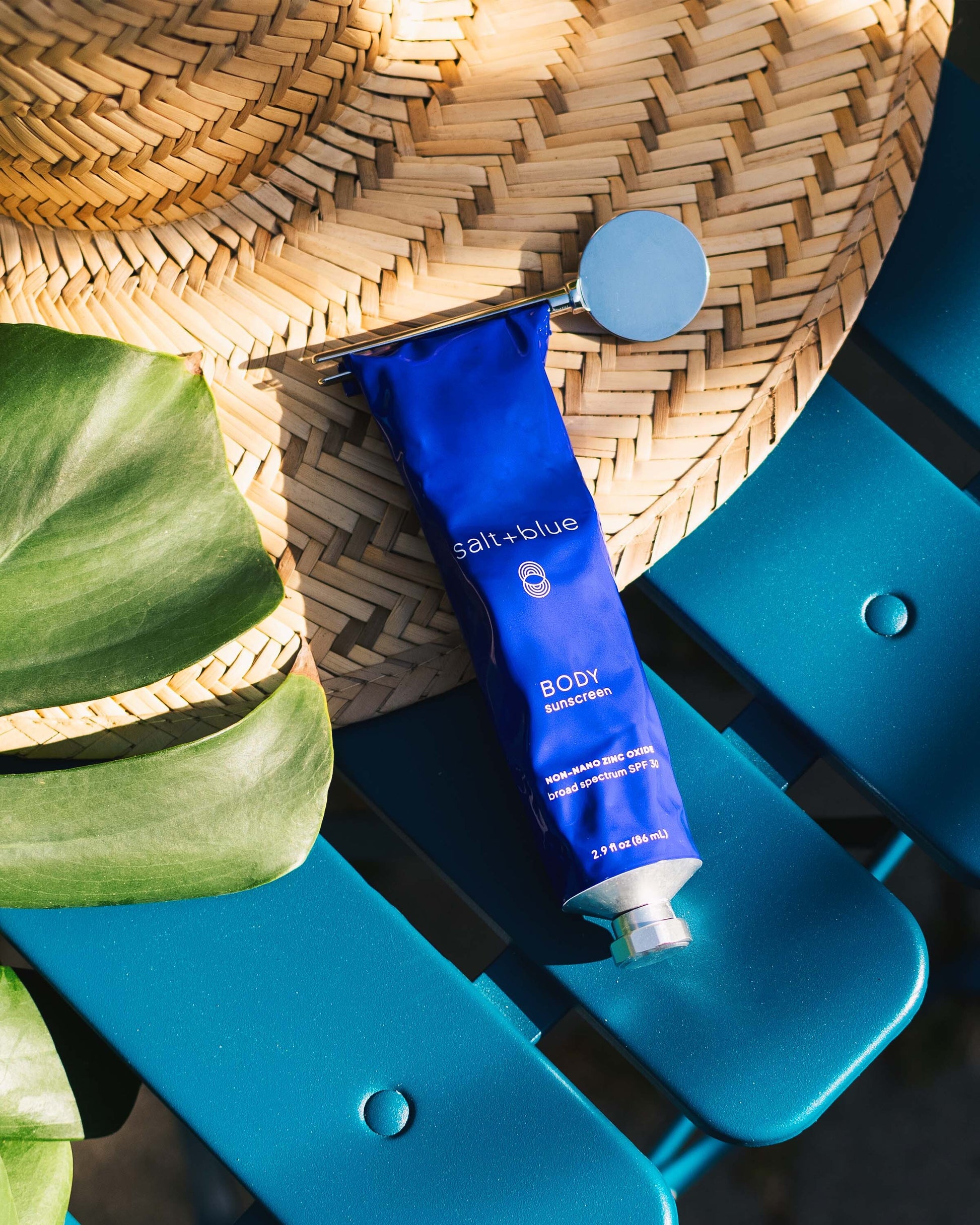 A tube of salt+blue BODY sunscreen in a vibrant blue aluminum tube with a tube key rolled at the top edge, styled on a woven straw hat and a bright teal picnic table with green leaves in the foreground.