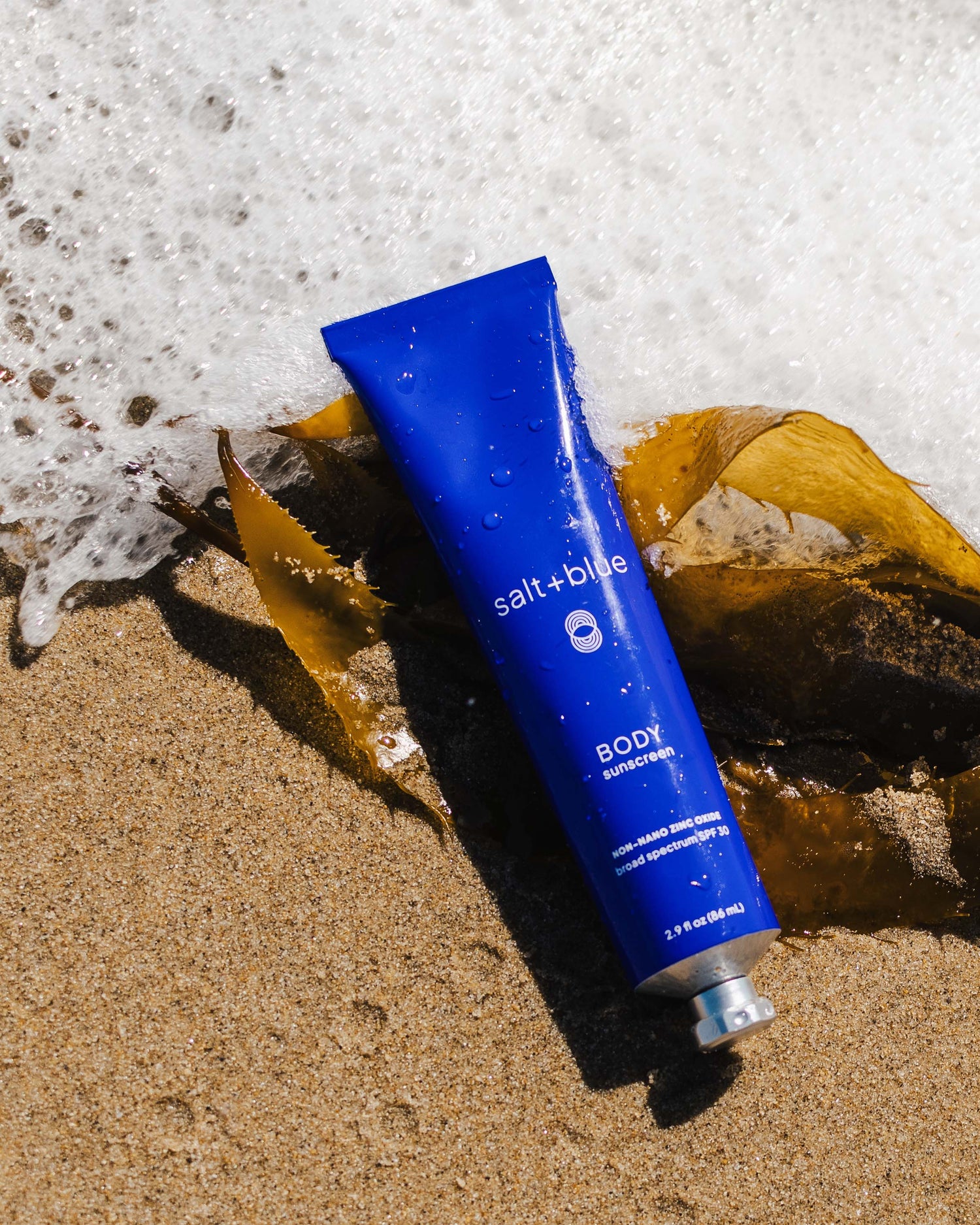 Blue tube of salt+blue body sunscreen on sand with seaweed and seafoam