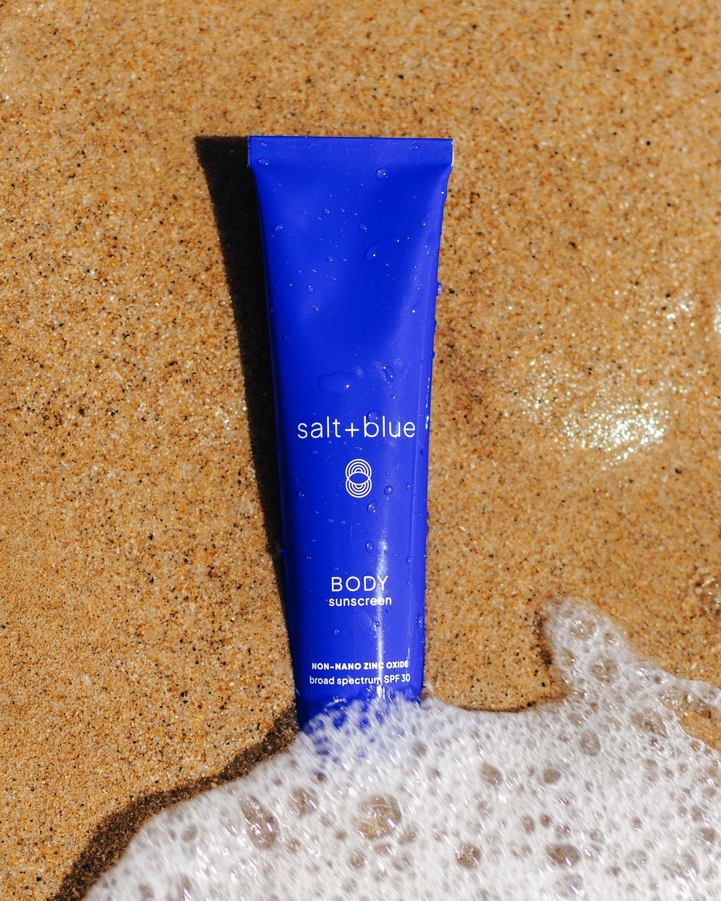 Blue sunscreen tube labeled 'salt+blue' on sandy beach with water.